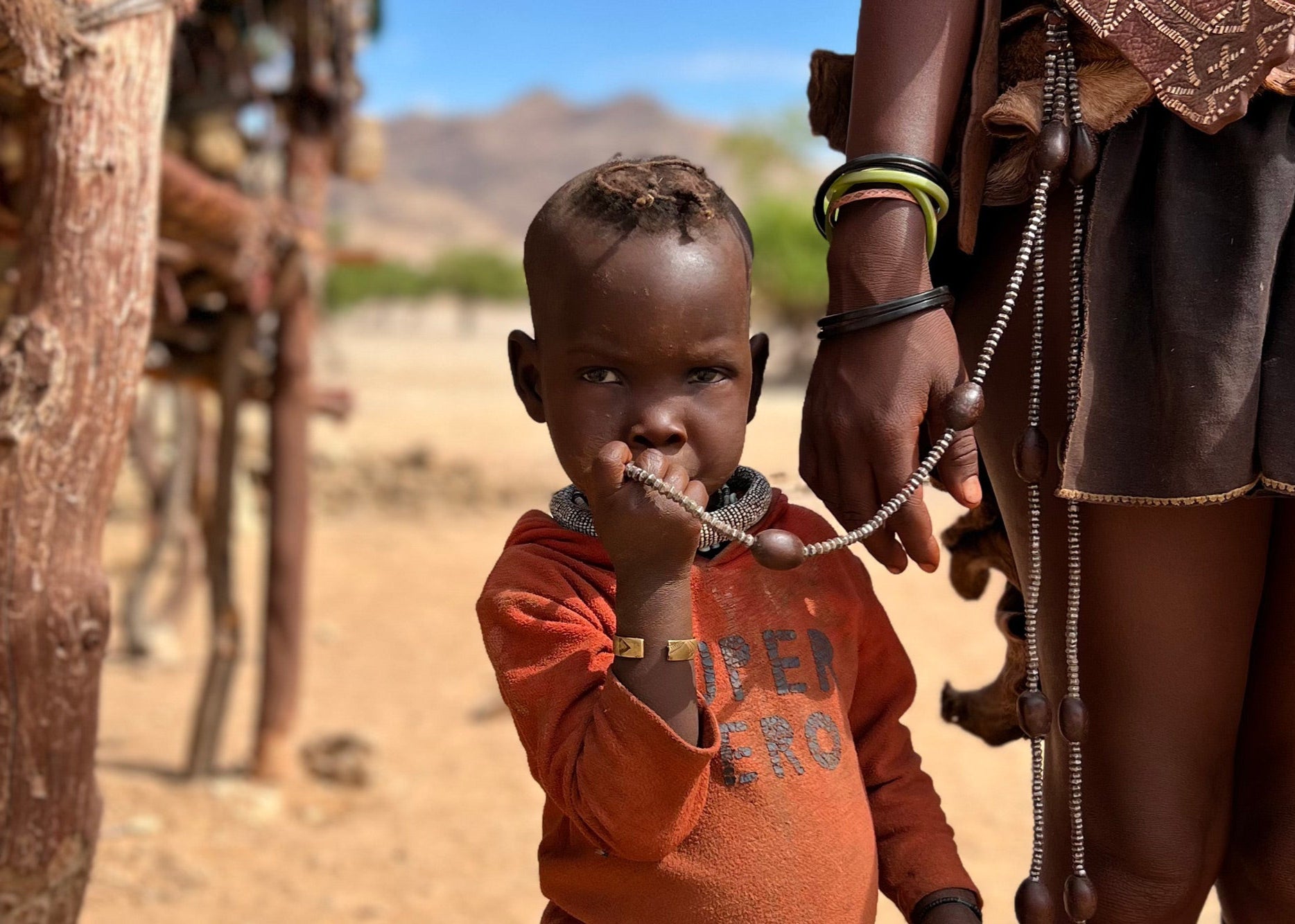 Child in an orange shirt standing next to a person in traditional attire in a desert-like setting.